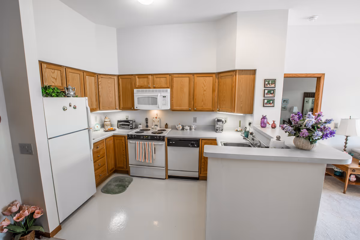 A bright kitchen with wooden cabinets, white countertops, and white appliances including a refrigerator, stove with oven, microwave, and dishwasher. There is a small green rug on the floor in front of the stove. A basket with purple flowers is placed on the countertop near the sink. The kitchen opens into a living area with a lamp and side table visible through a doorway.
