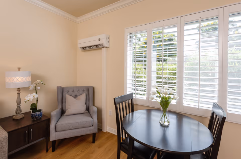 Sunlit dining nook with a round dark wood table and vase of flowers next to a gray upholstered armchair and side table.