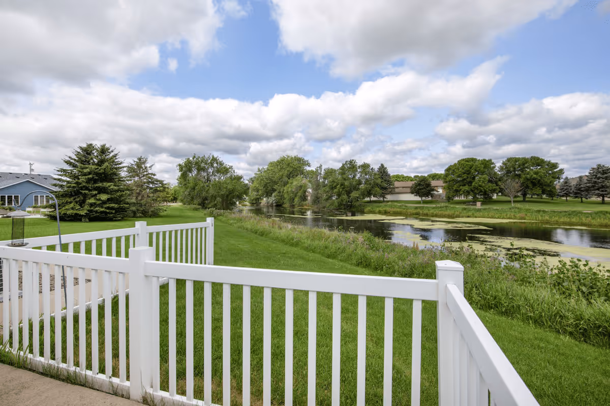 View of a peaceful outdoor area with a white fence in the foreground, green grass, trees, and a pond with water plants under a partly cloudy sky.