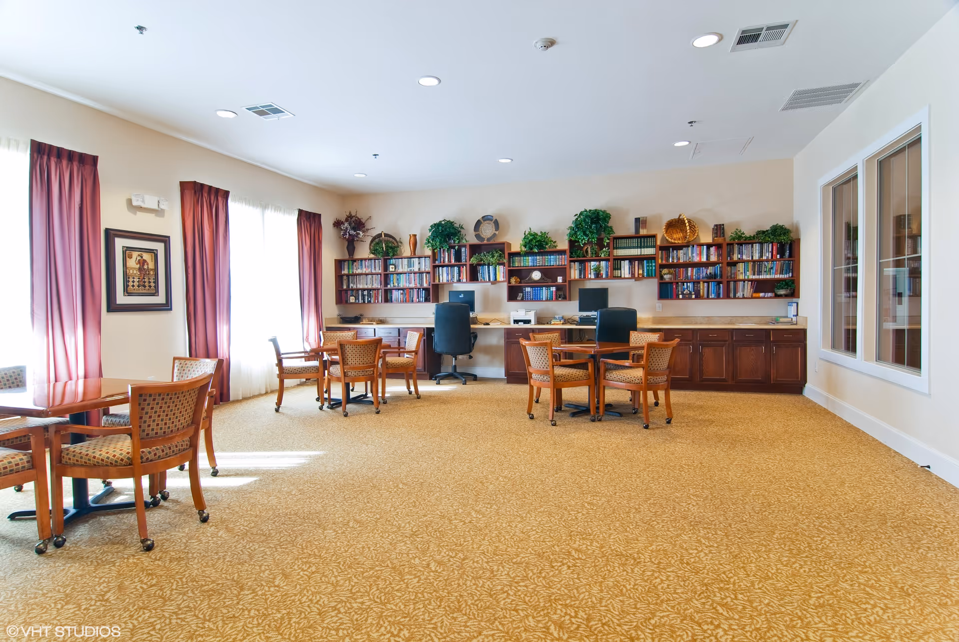 A bright and spacious common room with carpeted floor, several wooden tables and chairs, large windows with red curtains allowing natural light, and a wall lined with bookshelves filled with books and decorative plants. Two computer workstations with office chairs are also visible along the back wall.