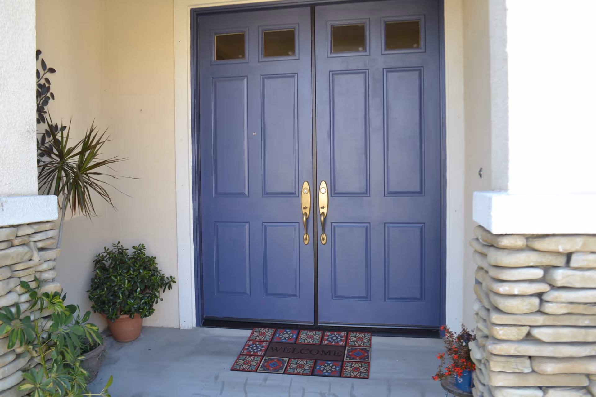 Front entrance with double blue doors featuring gold handles, a welcome mat with colorful patterns, and potted plants on either side under a covered porch with stone pillars.