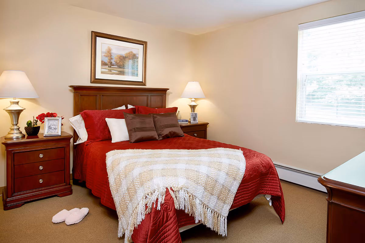 Cozy bedroom with red bedding, a wooden headboard, nightstands with lamps and decor, a framed picture above the bed, and a window with blinds.
