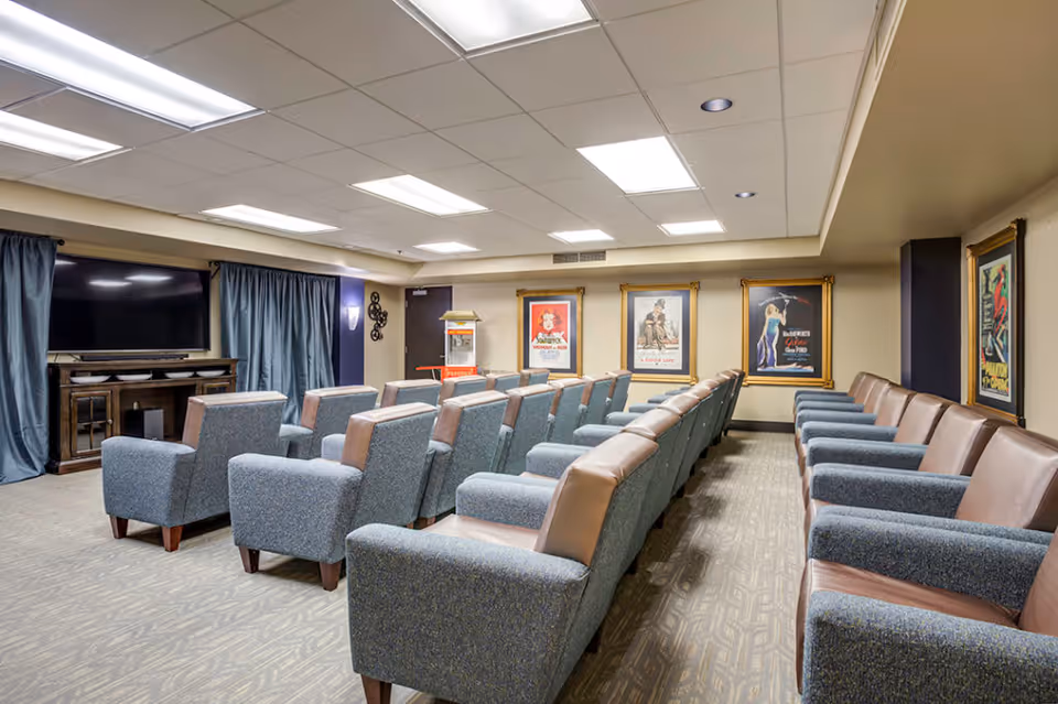 A cozy movie theater room with multiple rows of comfortable armchairs facing a large flat-screen TV mounted on a wooden cabinet. The walls are decorated with framed vintage movie posters, and there is a popcorn machine in the corner. The room has a carpeted floor and ceiling lights.
