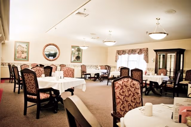 A dining room with multiple round tables covered with white tablecloths and surrounded by upholstered chairs with patterned fabric. The room has beige walls with framed artwork and a round mirror, carpeted floor, and ceiling lights. A window with a valance curtain is visible, along with a dark wood china cabinet in the corner.
