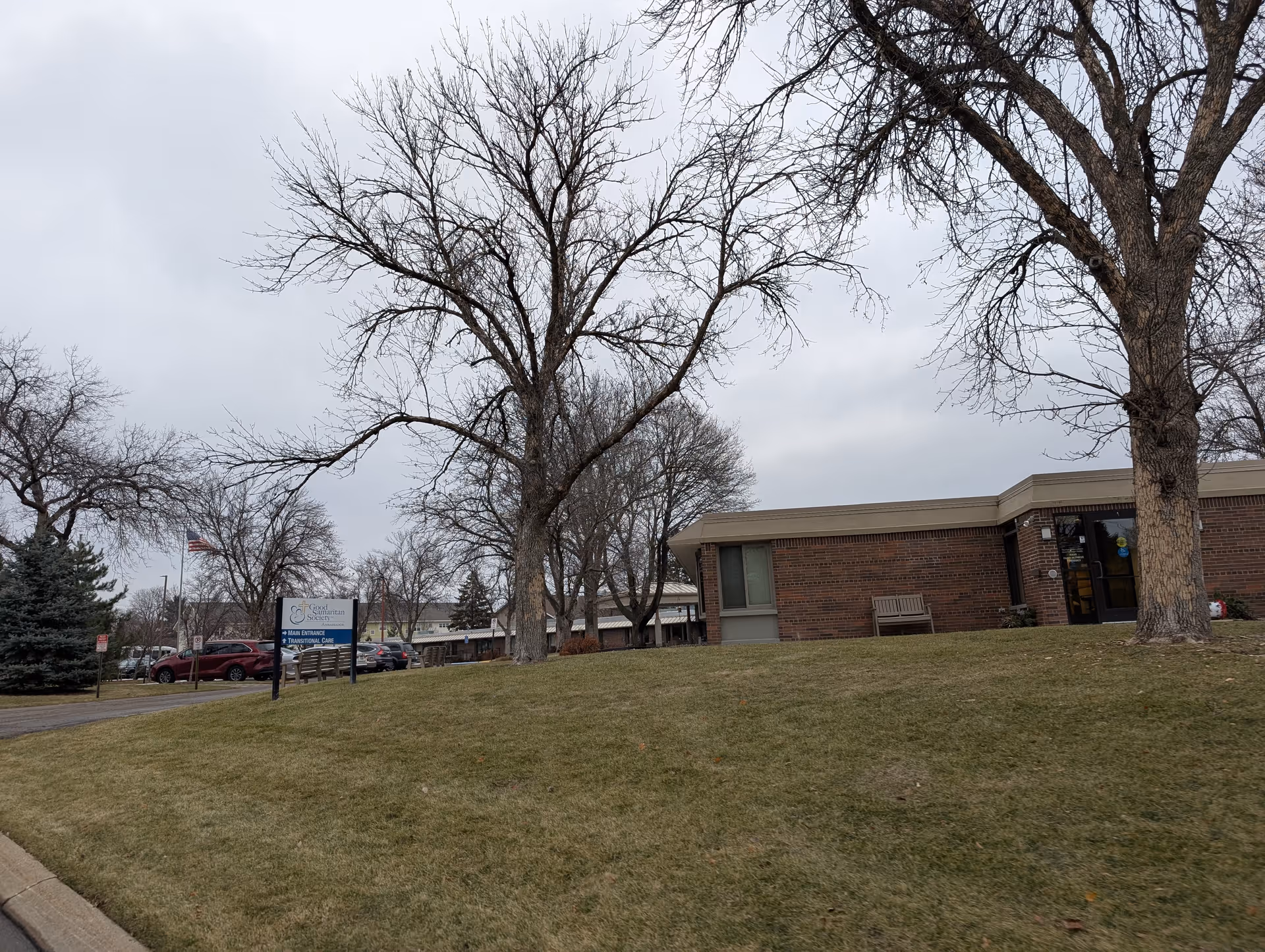 Exterior view of Good Samaritan Ambassador facility showing a single-story brick building with leafless trees and a grassy lawn in front. A sign near the parking lot indicates the main entrance and transitional care. Several parked cars and an American flag are visible in the background under a cloudy sky.