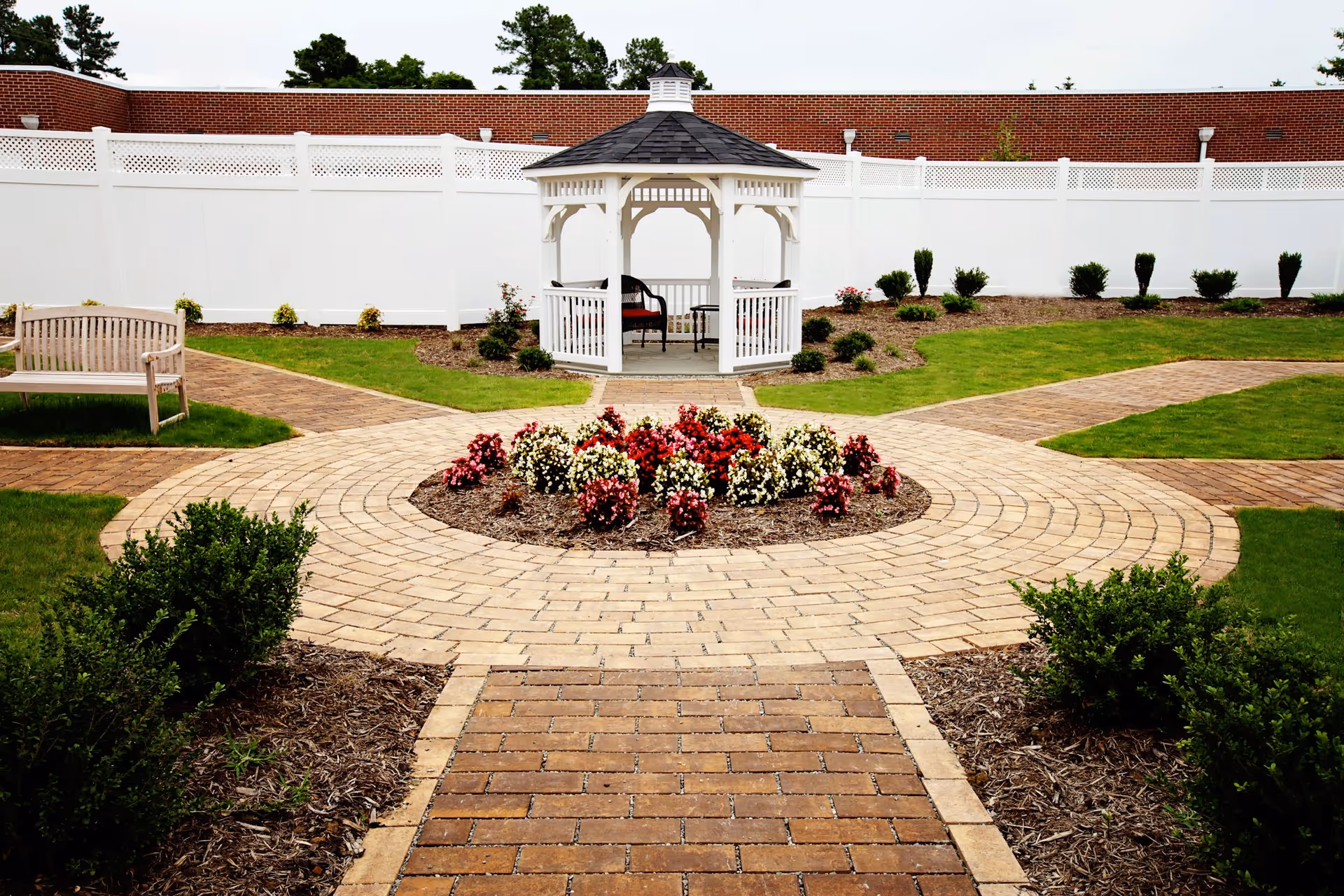 A landscaped outdoor garden area with a circular brick pathway surrounding a flower bed with red, white, and pink flowers. In the center background, there is a white gazebo with a dark shingled roof and seating inside. The garden is bordered by a white fence and a brick wall behind it, with green grass and small shrubs planted around the pathways.