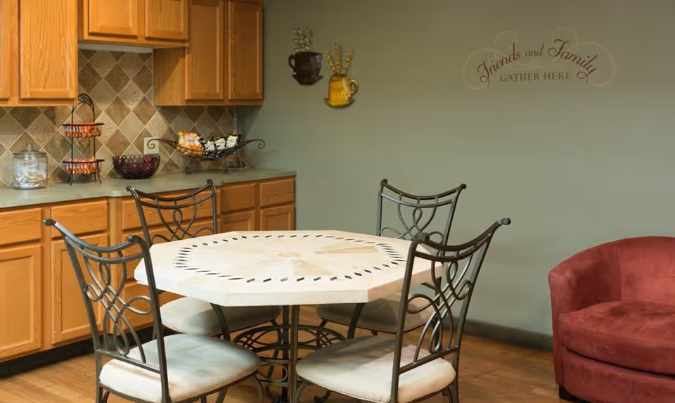 A cozy dining area with a round table surrounded by four metal chairs with cushioned seats. Behind the table, there is a kitchen counter with wooden cabinets and a tiled backsplash. On the counter, there are baskets and jars with snacks. The wall is painted green and has decorative wall art featuring two mugs and the phrase 'Friends and Family Gather Here'. A red upholstered chair is partially visible on the right side.