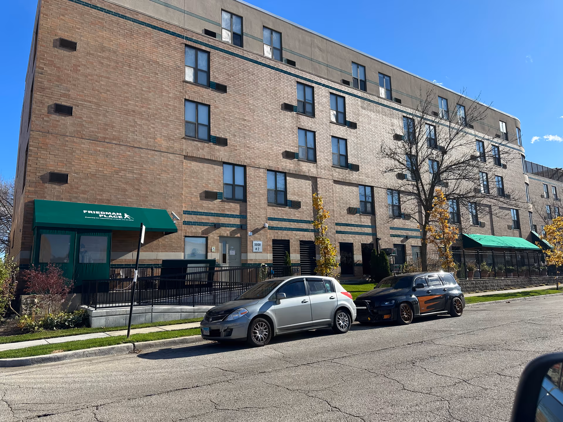Exterior view of a multi-story brick building with green awnings labeled 'Friedman Place'. Two cars are parked on the street in front of the building, and there are small trees and a sidewalk along the street. The sky is clear and blue.
