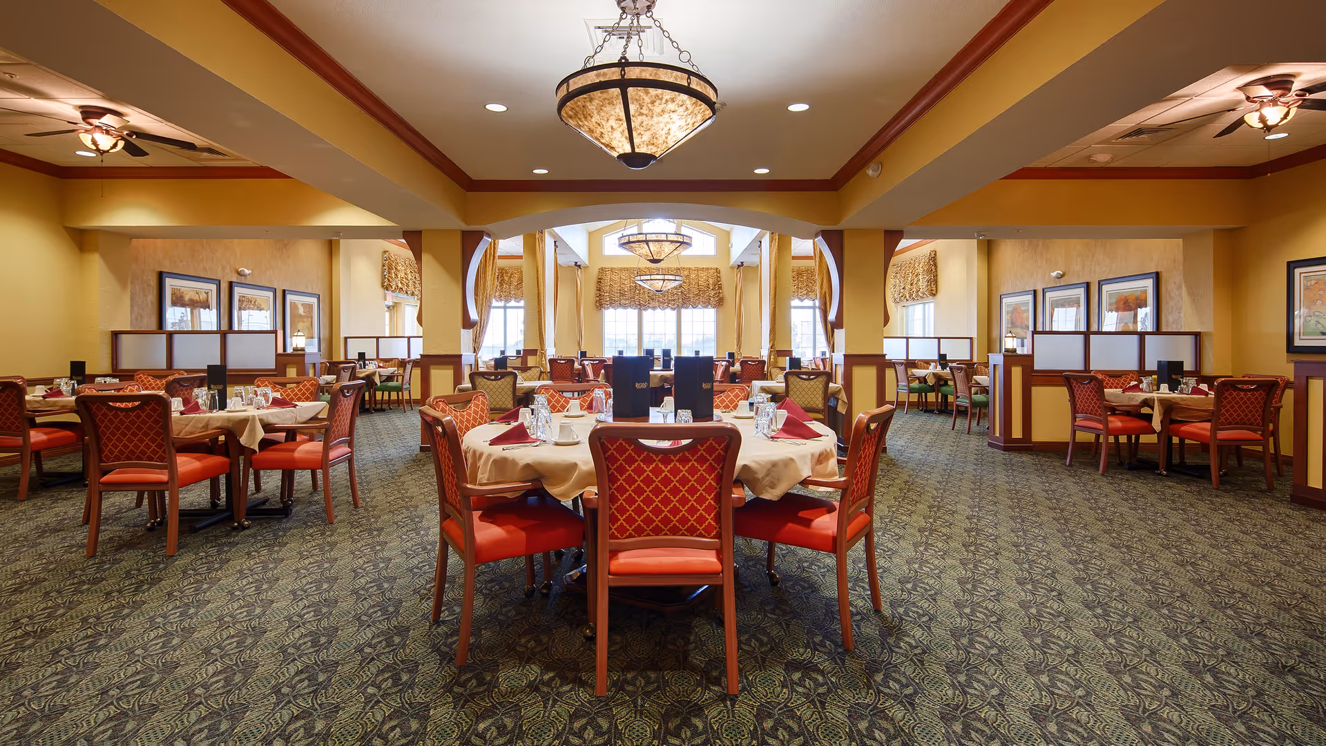 A spacious dining room with multiple round tables covered with beige tablecloths and set with glassware, napkins, and menus. The chairs have red cushioned seats and backs with a diamond pattern. The room features warm yellow walls, large windows with valances, framed artwork, and ceiling fans with lights. The carpet has a green and blue patterned design.