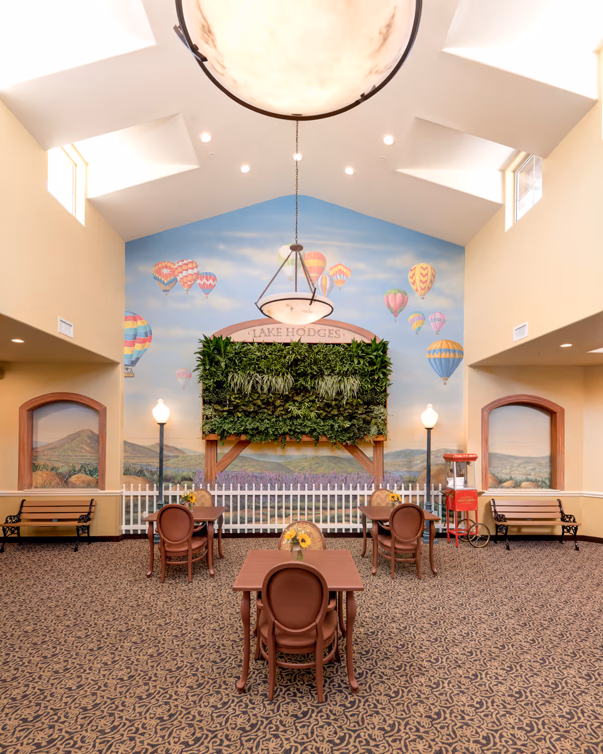 A bright and spacious common area with high ceilings and skylights. The room features a mural of hot air balloons in the sky above a landscape with mountains and fields. There is a vertical garden mounted on the wall with the sign 'Lake Hodges' above it. The room has several small tables with chairs, benches along the walls, a popcorn machine, and decorative street lamps. The carpet has an intricate pattern.
