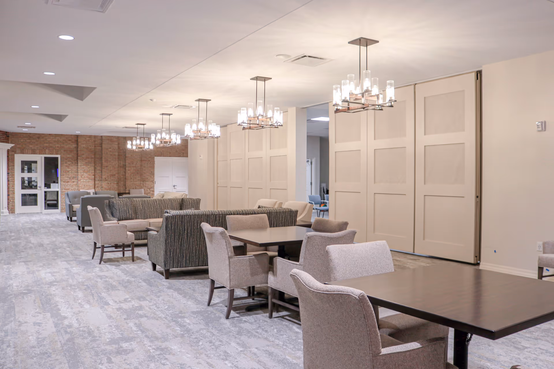 A spacious, well-lit common area with multiple seating arrangements including armchairs, sofas, and tables. The room features modern chandeliers hanging from the ceiling, a light-colored carpet, and a brick accent wall with glass doors at the far end.