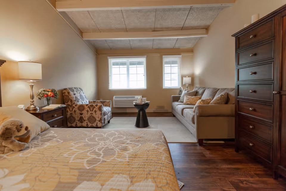 A cozy living space featuring a bed with a floral-patterned quilt and a stuffed animal, a wooden nightstand with a lamp and flowers, a patterned armchair, a small black round table, a beige sofa with cushions, two windows with blinds, and a tall wooden dresser on the right side.