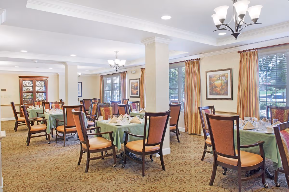 Bright dining room with round tables set for meals, upholstered chairs, chandeliers, and large windows with curtains.