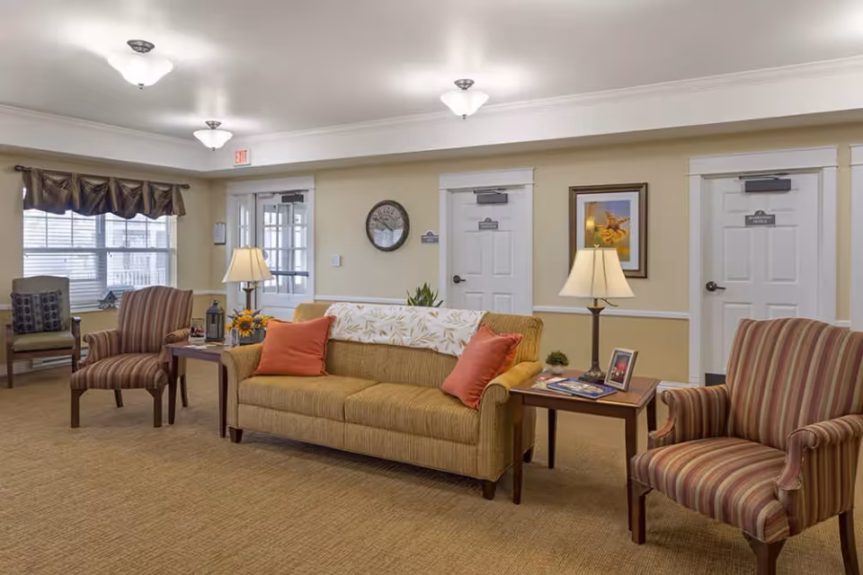 A cozy assisted living facility common area with a beige sofa adorned with two orange pillows and a white patterned throw. Two striped armchairs flank the sofa, each next to a wooden side table with lamps and decorative items. The room has beige walls, carpeted floor, three white doors, a clock, framed artwork, and a window with brown valance curtains letting in natural light.