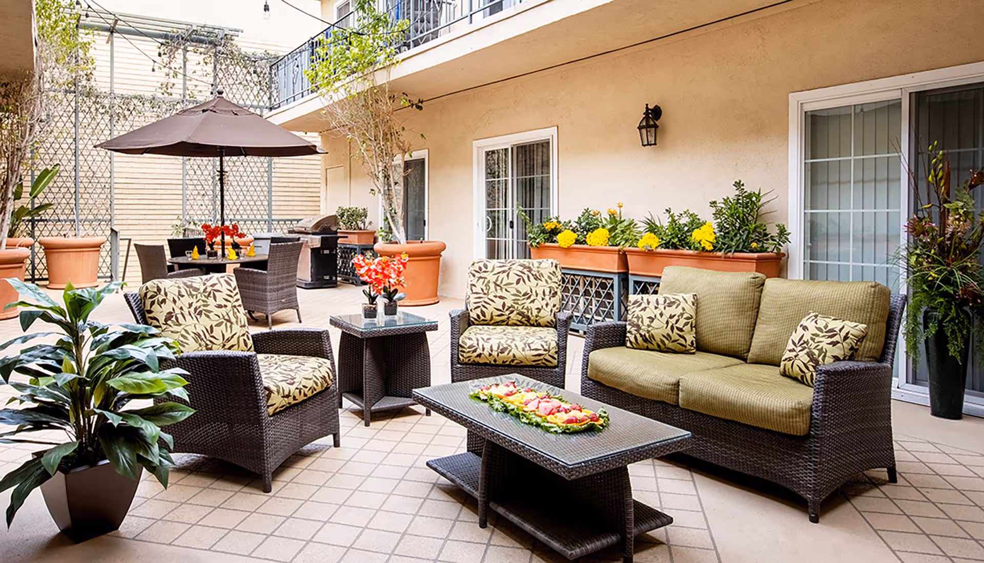 Outdoor patio area with wicker furniture including a sofa, two armchairs with leaf-patterned cushions, and a coffee table with a decorative fruit platter. There is a dining table with chairs and a large umbrella in the background, along with potted plants and flowers along the wall and a barbecue grill.