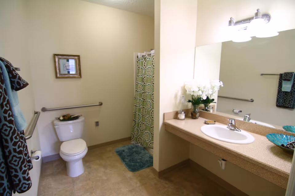 A clean and well-lit bathroom with a toilet, a shower area with a patterned curtain, a large mirror above a countertop with a sink, and decorative items including a vase with white flowers and a blue decorative bowl. Towels hang on the wall and a framed picture is above the toilet.