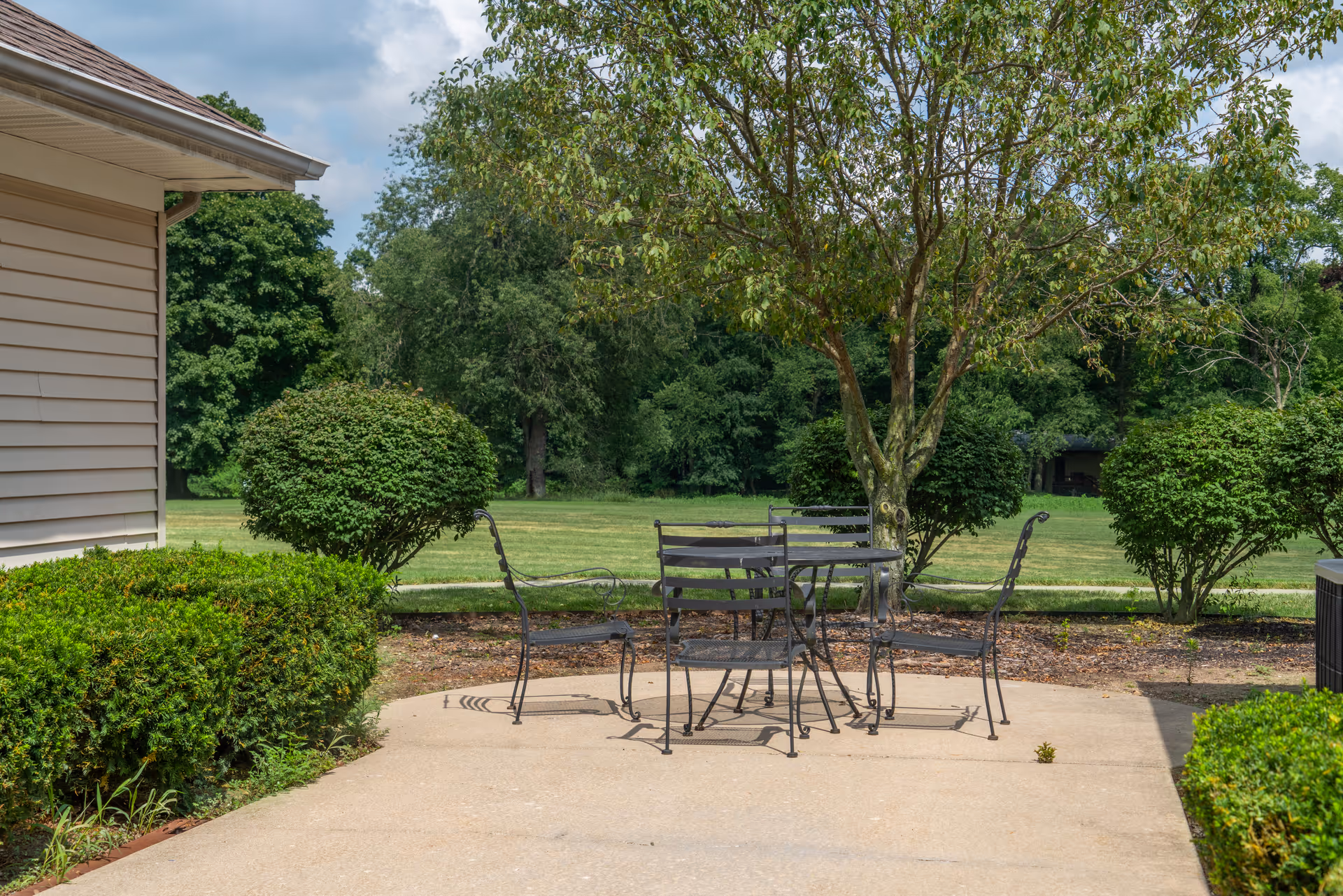 Outdoor patio area with a metal table and four chairs on a concrete surface, surrounded by green bushes and trees with a grassy field and more trees in the background under a partly cloudy sky.