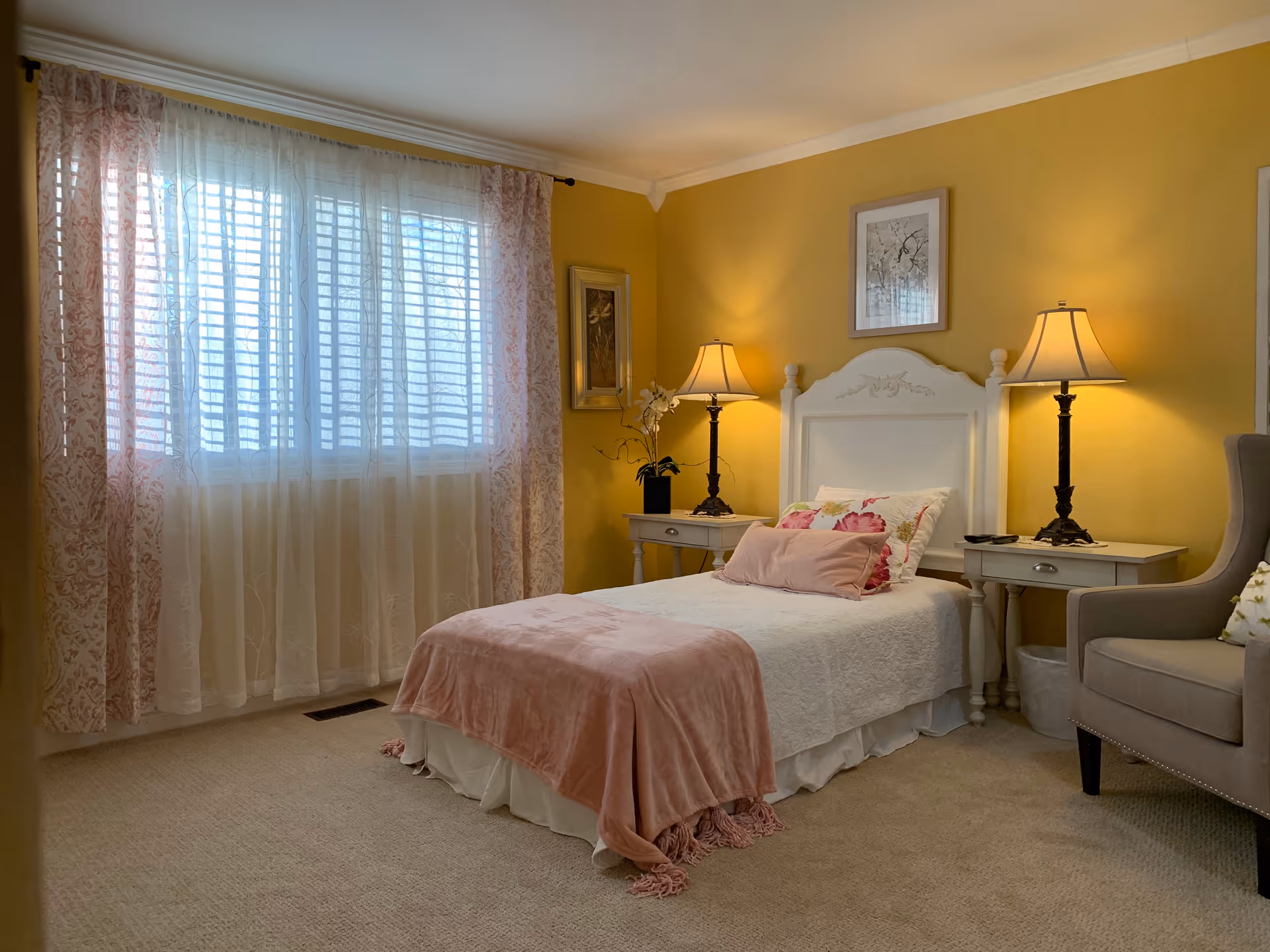 A cozy bedroom with a single bed covered in white bedding and a pink throw blanket. The bed has floral and pink pillows and a white headboard. On either side of the bed are matching white nightstands with lamps. A gray armchair is positioned to the right of the bed. The walls are painted yellow, and there is a window with sheer white and patterned pink curtains letting in natural light. Two framed pictures hang on the walls.