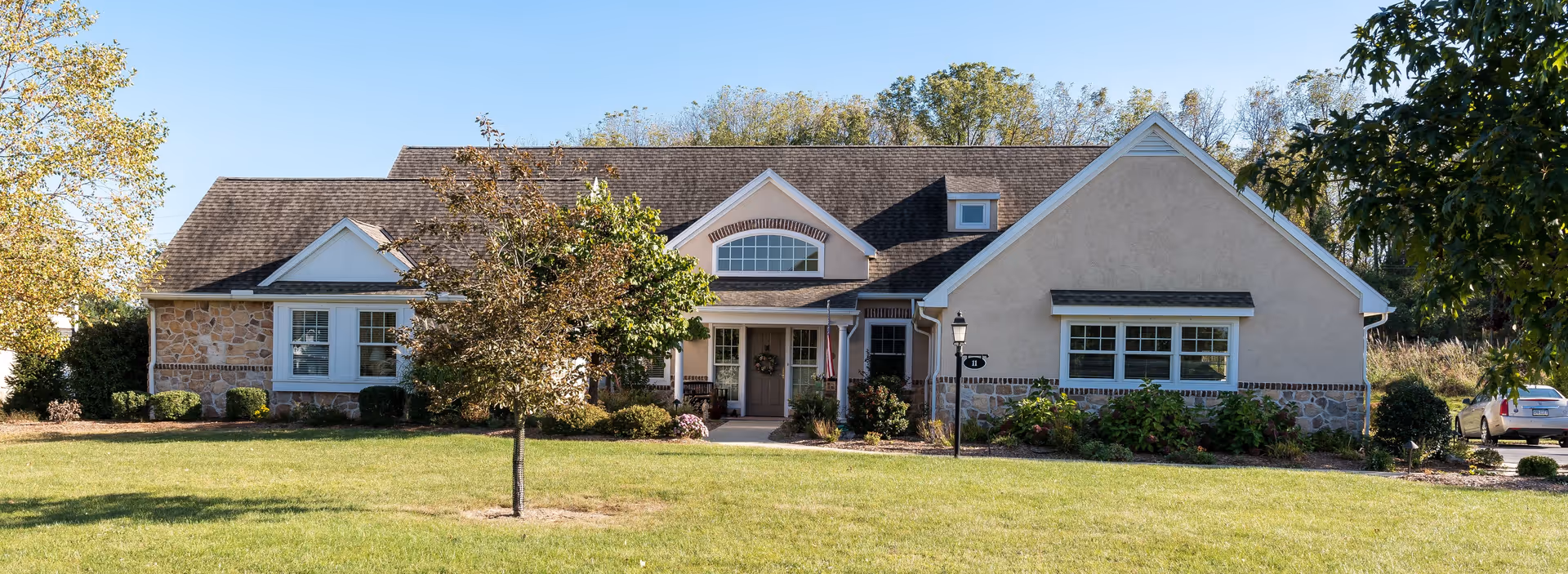 Front exterior view of a single-story residential building with a stone and stucco facade, multiple windows, a central entrance with a small porch, surrounded by a well-maintained lawn, trees, and shrubs under a clear blue sky.