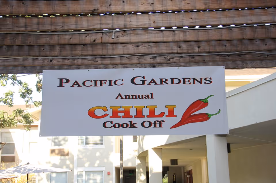 A banner reading "Pacific Gardens Annual CHILI Cook Off" hung beneath a wooden pergola in a facility courtyard.