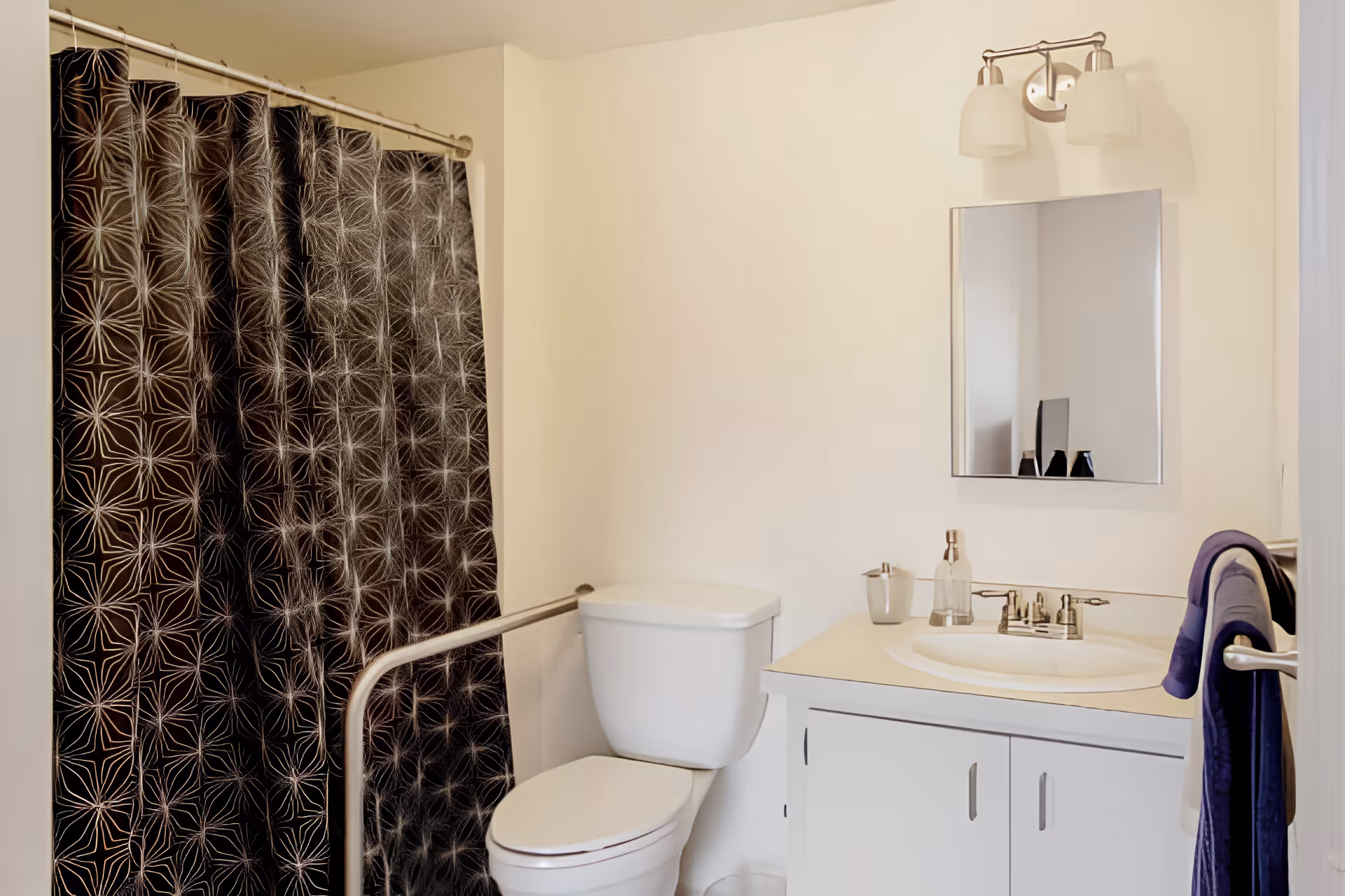 A clean bathroom with a toilet, sink and vanity, mirror, purple towel, and a shower with a black patterned curtain and grab bar.
