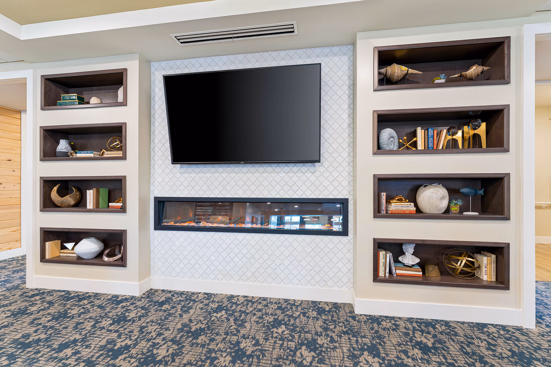 A modern interior wall featuring a large flat-screen TV mounted above a long, narrow electric fireplace. On either side of the TV and fireplace are built-in shelves with decorative items including books, sculptures, and ornamental objects. The floor is carpeted with a blue and beige patterned design.