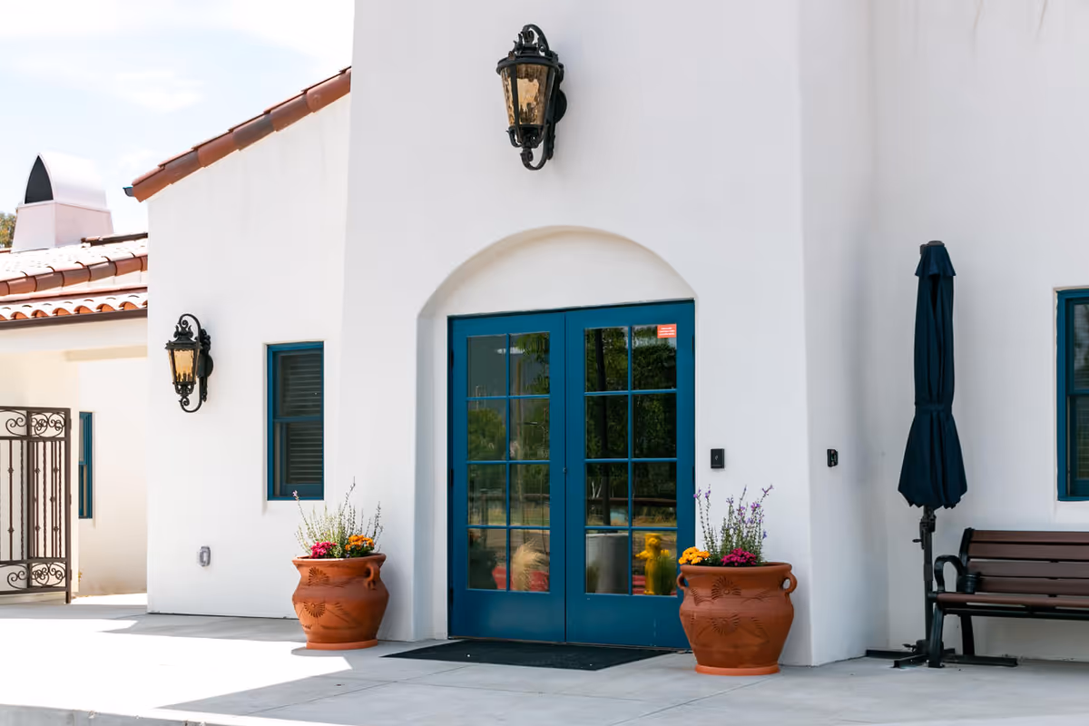 Exterior view of a white building with blue double doors, two large terracotta flower pots with colorful flowers on either side of the entrance, a black bench, a closed black umbrella, and decorative wall-mounted lanterns.