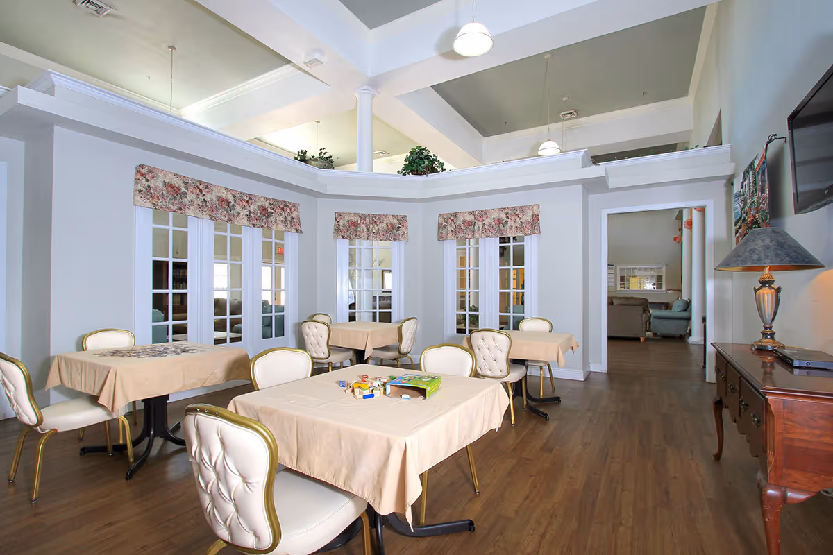 A bright and spacious dining area with several tables covered in beige tablecloths and surrounded by cushioned chairs. The room features large windows with floral valances, wooden flooring, and a high ceiling with hanging pendant lights. A wooden side table with a lamp and a wall-mounted TV are visible on the right side. In the background, there is an open doorway leading to a lounge area with sofas.