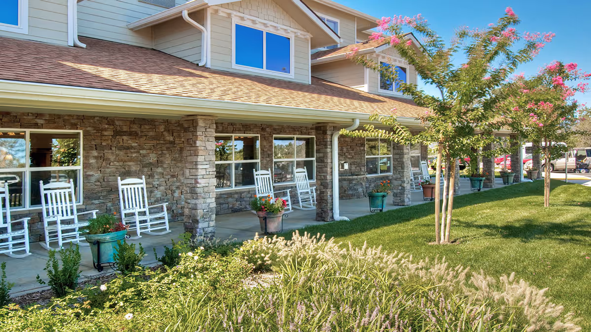 Covered front porch of a senior living building with white rocking chairs, potted plants, and a manicured lawn.