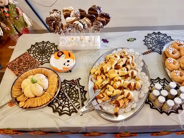 A table decorated with Halloween-themed items including black spider web placemats and small plastic spiders. On the table are trays of snacks: a plate with round crackers and a pumpkin-shaped cheese ball, a tray of crescent roll wrapped sausages resembling mummies, a plate of donuts decorated with eyes, and a stand holding chocolate-covered marshmallow pops. Small bottles of drinkable yogurt are also on the table.
