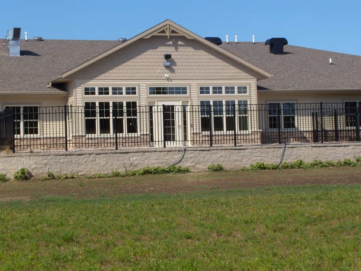 Exterior view of a single-story building with beige siding, multiple windows, and a central double door. The building is surrounded by a black metal fence and a low stone wall, with a grassy area in the foreground under a clear blue sky.