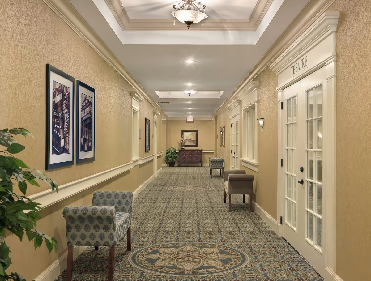 A well-lit hallway in a senior living facility with patterned carpet, beige walls, decorative ceiling lights, framed artwork on the walls, and several upholstered chairs along the sides. On the right side, there is a set of double doors labeled 'THEATRE'.