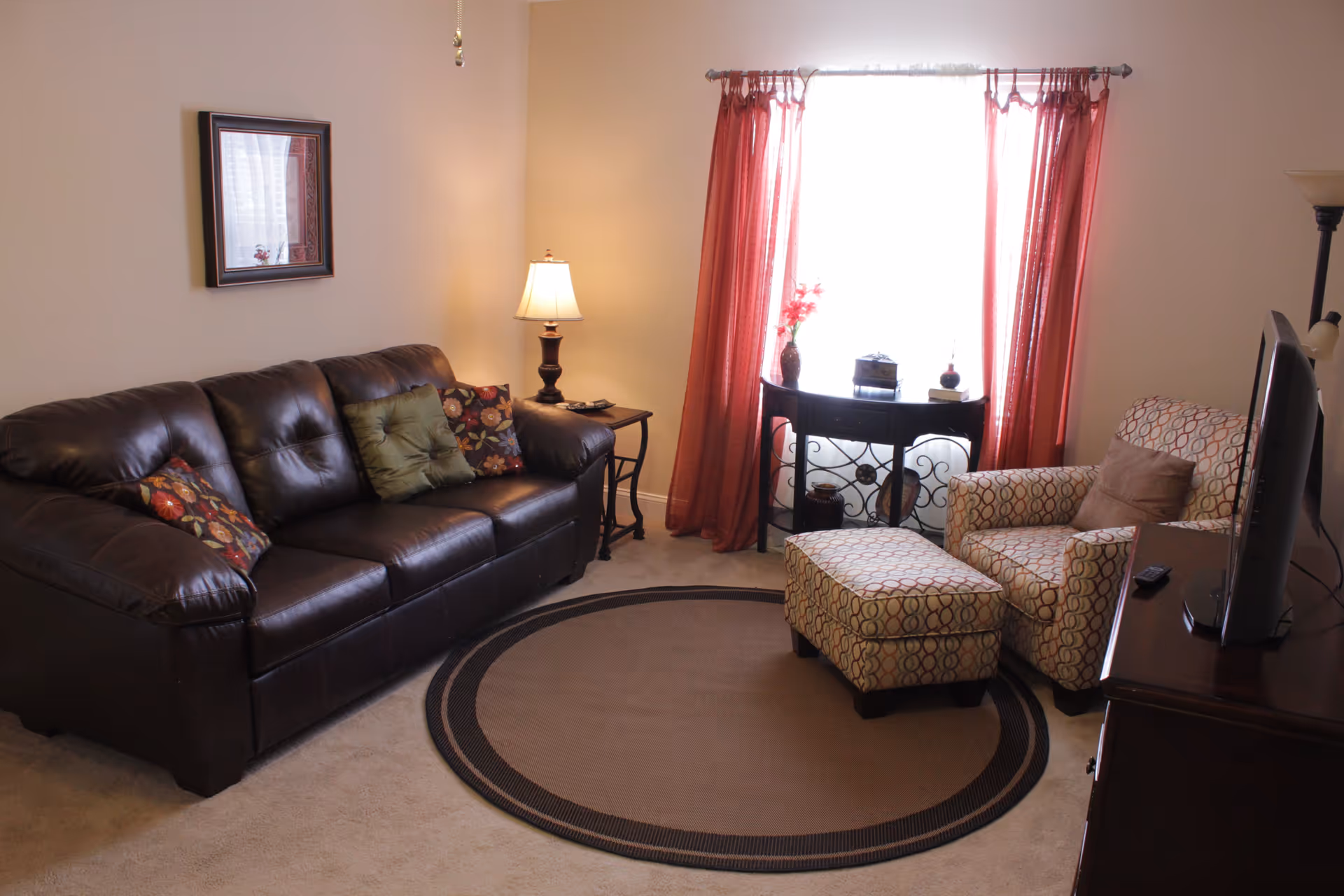 A cozy living room with a dark brown leather sofa adorned with colorful cushions, a patterned armchair with matching ottoman, a round area rug, a wooden side table with a lamp, a small console table with decorative items in front of a window with red curtains, and a flat-screen TV on a wooden stand.