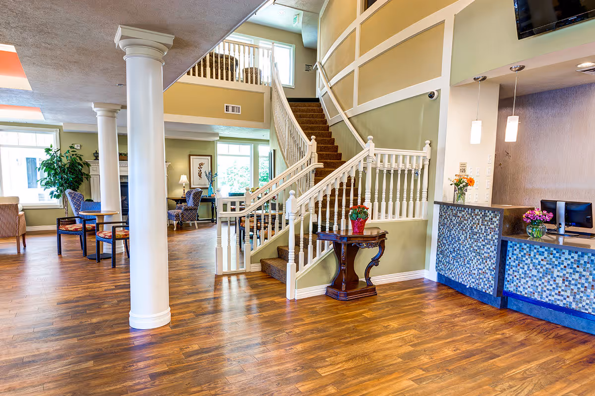 Bright senior living lobby with a curved staircase, reception desk, seating area, and hardwood floors.
