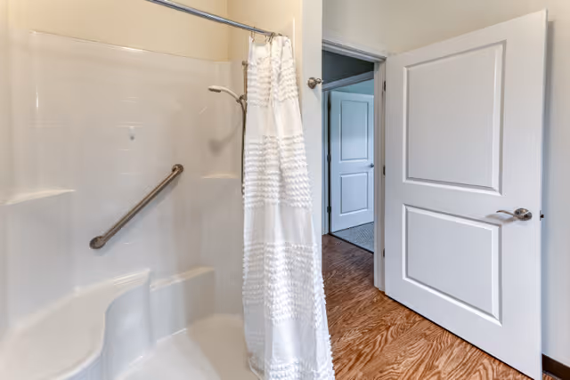 A bathroom with a white shower area featuring a built-in seat and a grab bar. A white shower curtain hangs on a curved rod. The bathroom door is open, showing a hallway with wooden flooring and another door in the background.