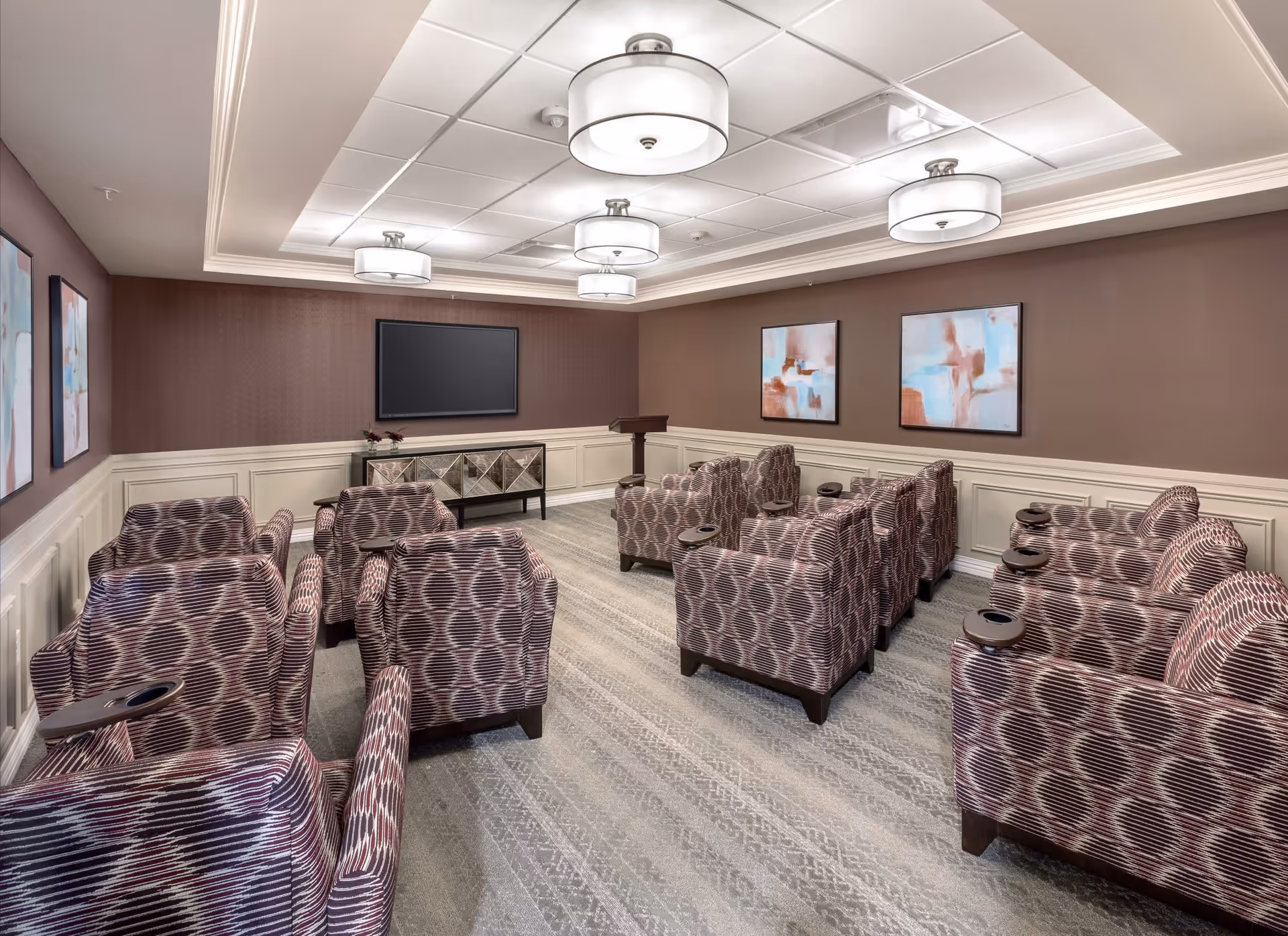 A small theater-style room with multiple patterned armchairs arranged in rows facing a large flat-screen TV mounted on a brown wall. The room has a light gray carpet, white wainscoting, abstract artwork on the walls, and four ceiling light fixtures.