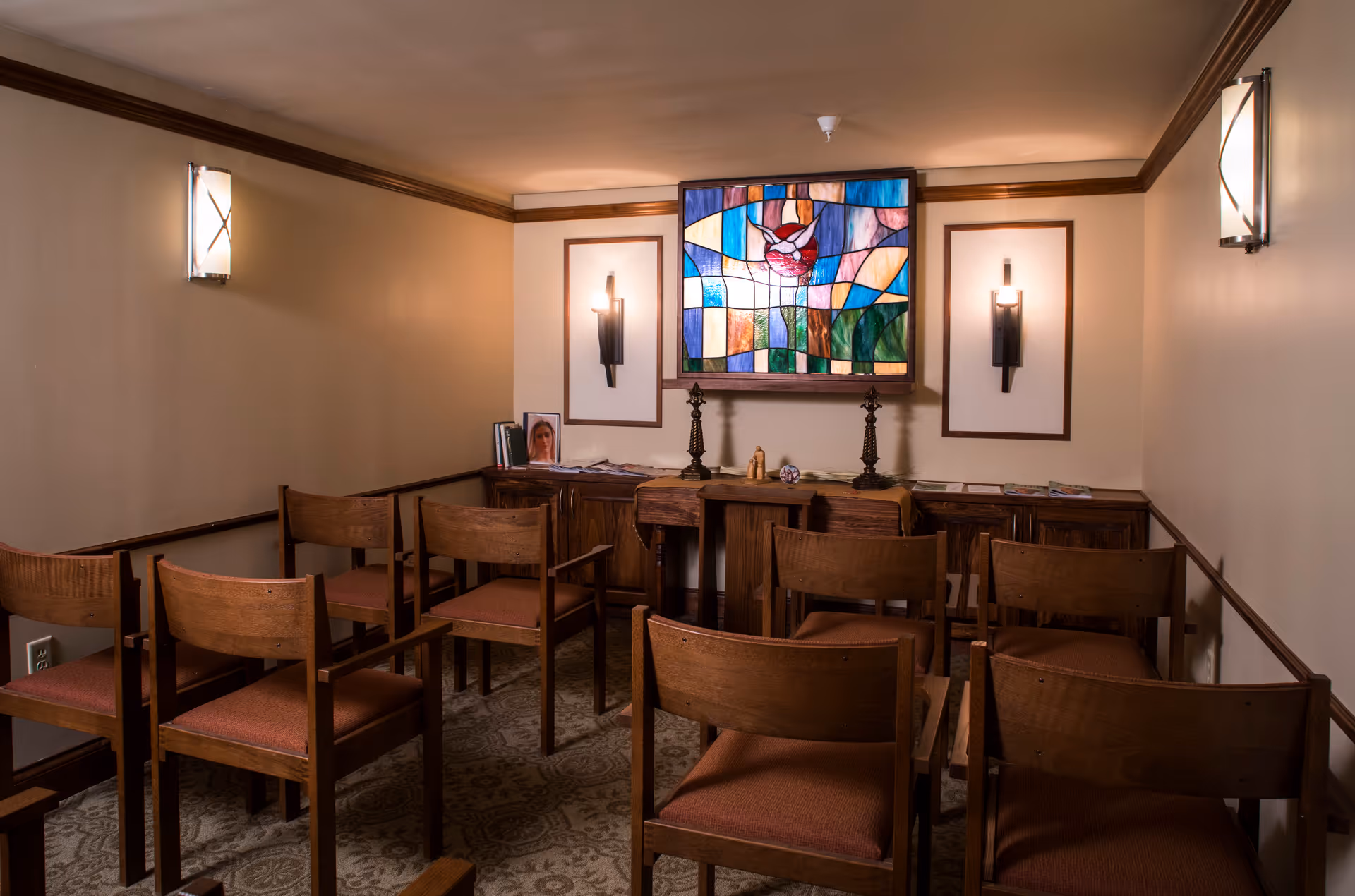 Small room with wooden chairs arranged in rows facing a wooden table against the wall. Above the table is a colorful stained glass artwork depicting a dove. The room has beige walls with wooden trim and wall-mounted lights on either side of the stained glass.