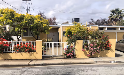 A yellow care home with a white gate and blooming roses along a sidewalk.