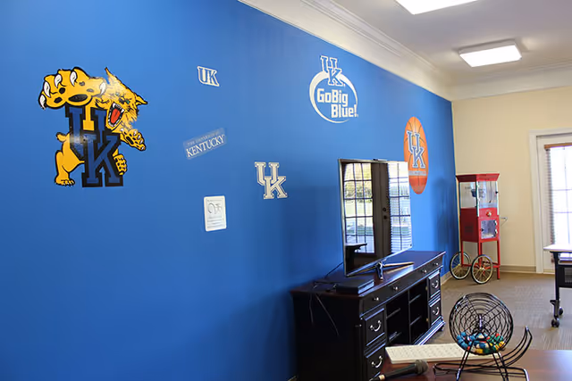 A room with a blue wall decorated with University of Kentucky logos and slogans. A flat-screen TV sits on a dark wooden cabinet below the logos. To the right, there is a red popcorn machine and a window with blinds. In the foreground, there is a bingo cage with colorful balls and a microphone on a table.