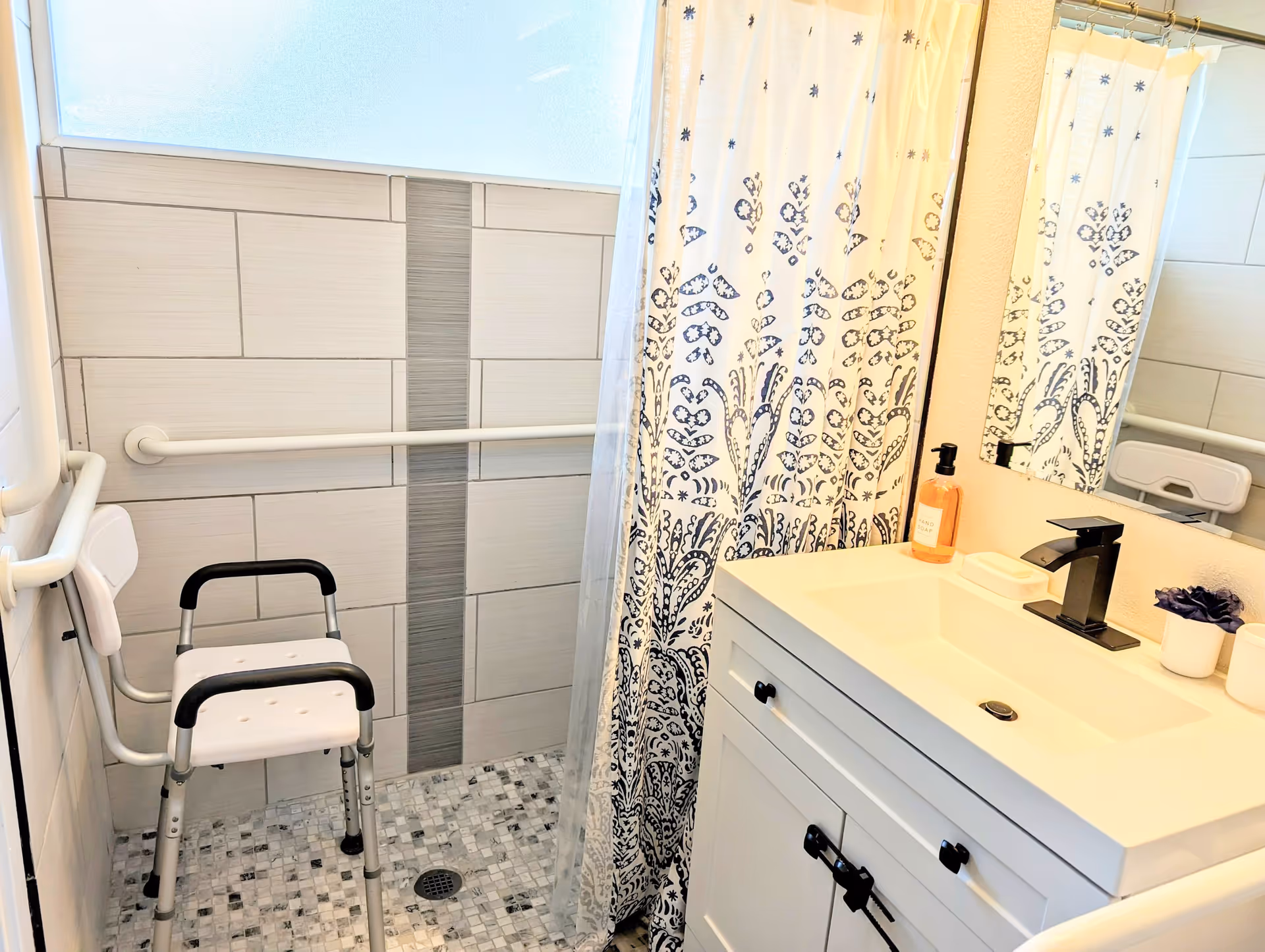 A bathroom with a walk-in shower featuring a white shower chair with black handles, grab bars on the tiled walls, a shower curtain with a blue and white pattern, and a white vanity with a black faucet, soap dispenser, and small potted plant.