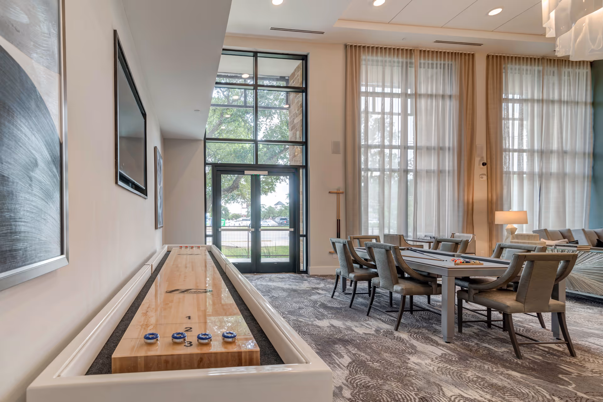A bright and spacious game room with a shuffleboard table in the foreground and a pool table surrounded by chairs in the background. Large windows with sheer curtains allow natural light to fill the room. The room has modern decor with a patterned carpet and a wall-mounted TV.