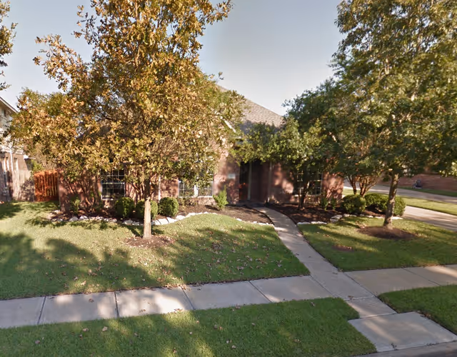 Front exterior view of a single-story brick building partially obscured by trees and bushes, with a concrete walkway leading to the entrance and a well-maintained lawn.