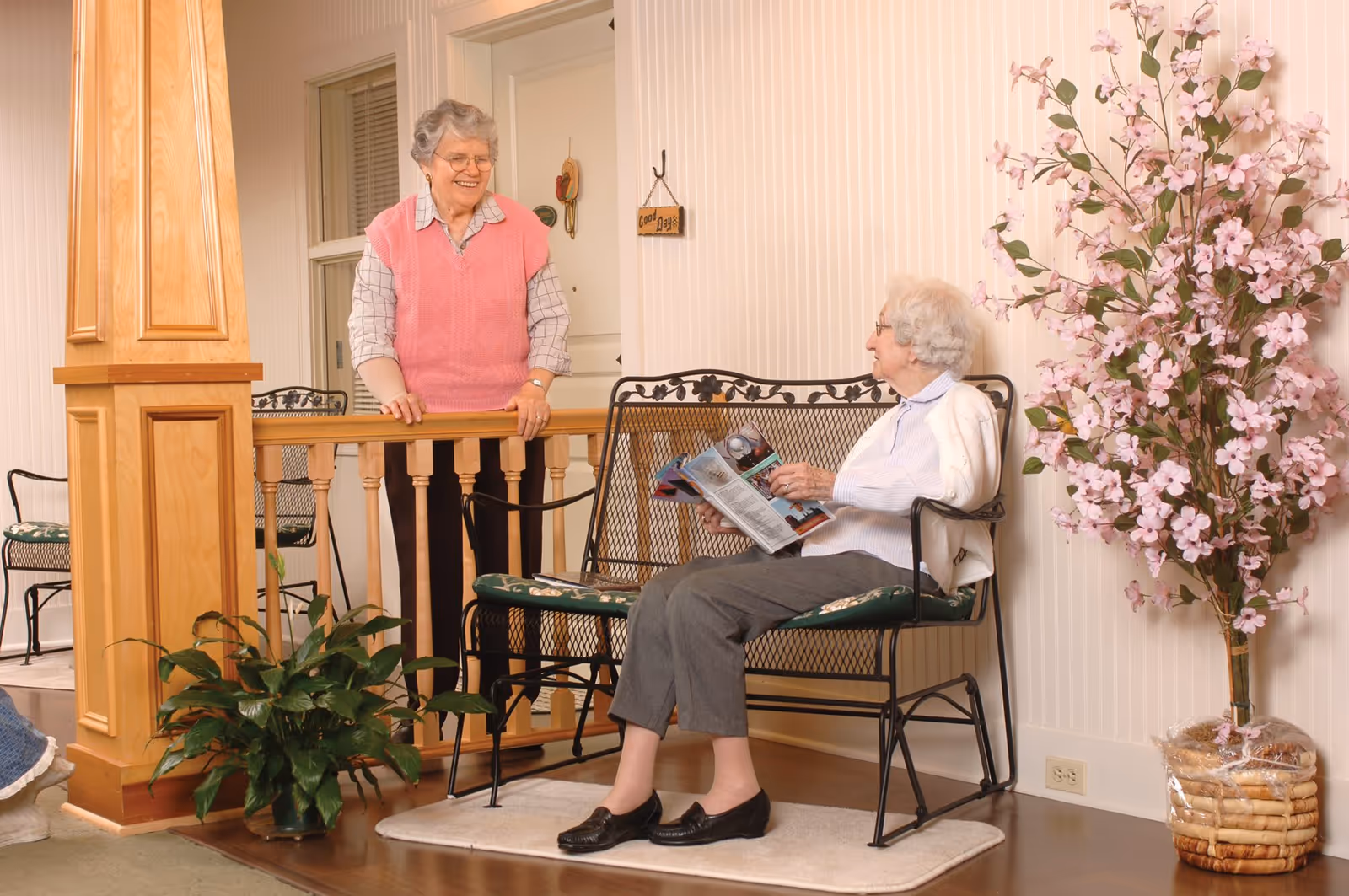 Two elderly women in a cozy indoor setting; one woman with gray hair and glasses wearing a pink sweater vest is standing and smiling while leaning on a wooden railing, and the other woman with white hair and glasses is sitting on a black metal bench with green cushions, reading a magazine. There is a large potted plant with pink flowers on the right and a green leafy plant near the wooden column on the left.