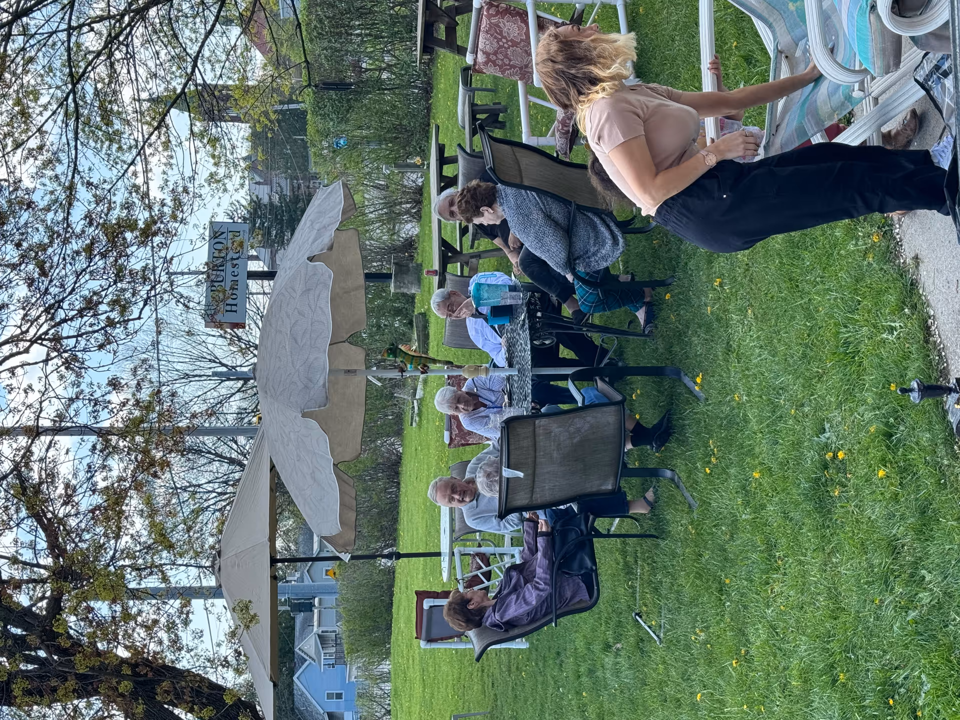 A group of elderly people sitting around a round outdoor table with umbrellas in a grassy yard, engaged in conversation. A younger woman is bending over near a lounge chair in the foreground. Trees and bushes surround the area, and a sign in the background reads 'Burton Homestead'.
