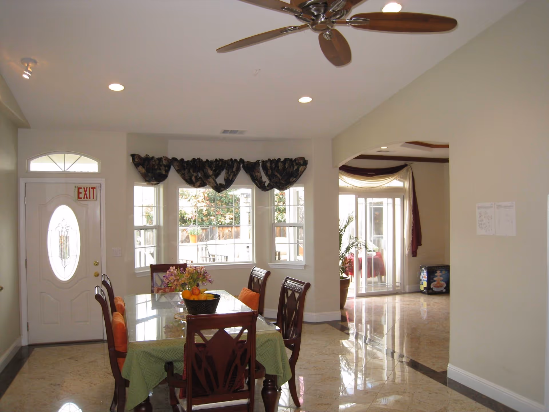 Interior view of a dining area with a glass-top table covered by a green tablecloth and surrounded by six wooden chairs with orange cushions. A basket with fruit and a flower arrangement are on the table. The room has beige walls, a ceiling fan with wooden blades, recessed lighting, and a white door with an exit sign above it. Large windows with dark floral valances let in natural light, and a sliding glass door leads to another room with a potted plant visible.