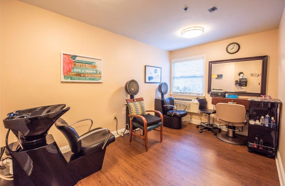 A small hair salon room with a black hair washing station and chair on the left, two vintage hair dryers with chairs in the middle, and a styling station with a large mirror, chair, and various hair products on the right. The room has light beige walls, a wooden floor, a window with blinds, and a clock above the mirror.