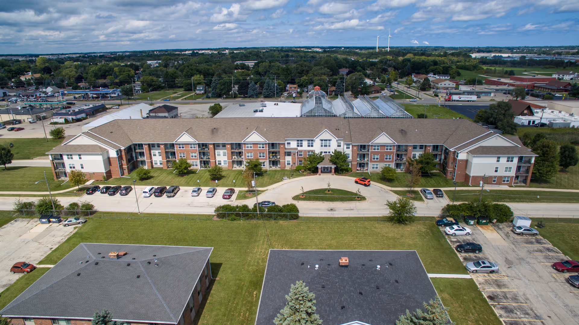 Aerial view of a senior living facility named Killarney Kourt, showing a large three-story building with a circular driveway and parked cars in front. The building is surrounded by green lawns and additional smaller buildings, with a suburban area and trees in the background under a partly cloudy sky.