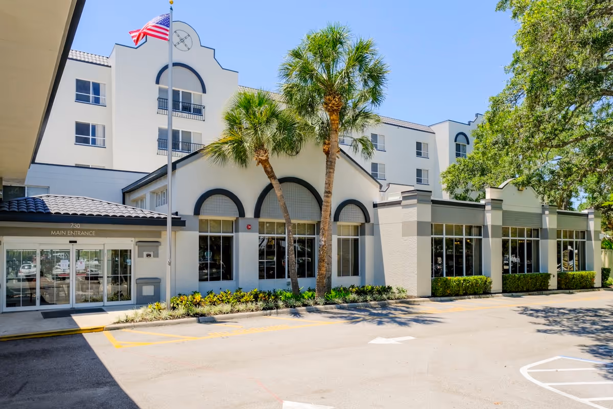 Exterior view of the Grand Villa of Sarasota building with a main entrance, palm trees, an American flag on a flagpole, and a clear blue sky.
