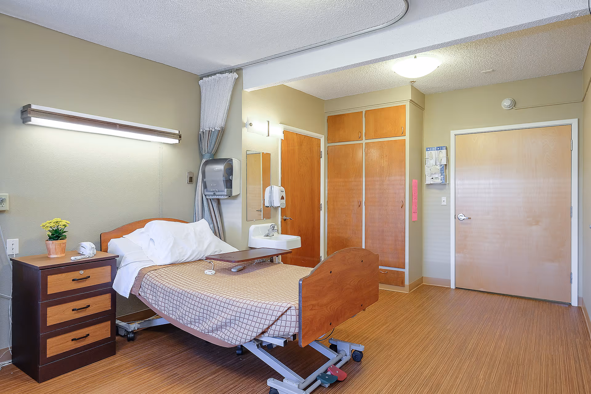 A clean, well-lit patient bedroom with a hospital bed, nightstand, sink, and wooden cabinets.