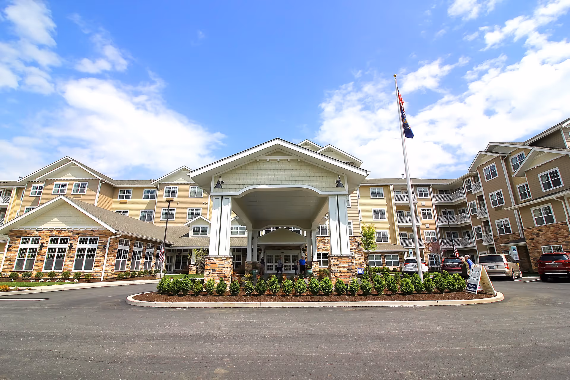 Front exterior view of Ivy Creek Gracious Retirement Living facility with a large covered entrance, multiple windows, and a flagpole in front. Several cars are parked near the entrance under a partly cloudy blue sky.